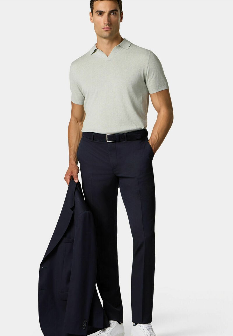 Světle šedý polo triko s krátkým rukávem, námořnické kalhoty, držící tmavý oblekový sako, doplněné bílými teniskami, se štíhlým střihem.
