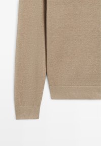 Manche et ourlet inférieur d'un pull en maille beige avec des poignets côtelés, présentés sur un fond blanc.