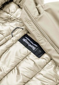 Giacca puffer beige con texture trapuntata, dotata di zip e di un'etichetta con il nome del marchio. L'ovatta interna è liscia e lucida.