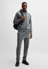 Grijze trainingsset van zacht materiaal; hoodie met ritssluiting en zwarte accenten en logo, gecombineerd met slim-fit broek en zwarte sneakers.
