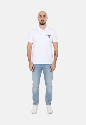 Uomo con barba e tatuaggio indossa una polo bianca, jeans azzurri e sneakers bianche, in piedi davanti a uno sfondo semplice.