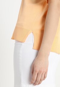 Persoon in een perzikkleurige top met korte mouwen en zijsplitten, met witte broek, zichtbaar vanaf de taille tot de hand die op de dij rust.