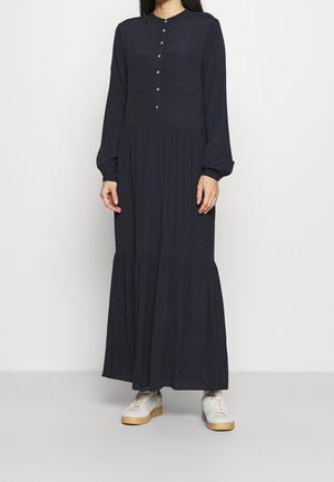 Kvinde iført en langærmet, marineblå maxi-kjole med knapper ned over brystet, kombineret med hvide sneakers med gummisåler.