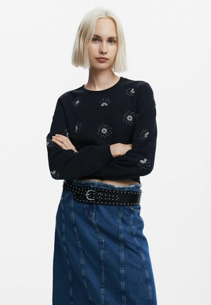 Felpa cropped blu navy con ricami floreali in argento. Abbinata a una gonna di jeans a vita alta con orlo sfrangiato e cintura con borchie.