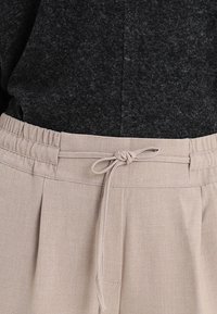 Beige byxor med strukturerad yta, som har dragsko i midjan och veckdesign. Tyget verkar mjukt och lätt.