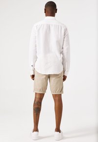 Chemise en lin blanche à manches longues avec des poignets boutonnés, associée à un short beige retroussé. Baskets blanches et un tatouage visible sur la jambe.