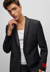 Čierny blazer so saténovými klopami, jedným náprsním vreckom a červenou nálepkou s nápisom "HUGO." Nosený cez biele tričko bez rukávov. Hladká textúra látky.