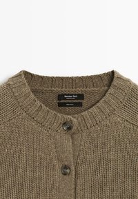 Bruine gebreide wollen cardigan met een ronde hals, voorzien van twee zwarte knopen en een gestructureerd patroon. Merkenlabel zichtbaar aan de binnenkant.