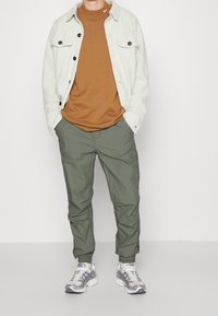 Ljusgrå fleecejacka med svarta knappar, brun crew neck t-shirt, gröna tapered byxor och silverfärgade löparskor med vita detaljer.