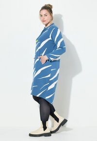 Blauwe langemouwen jurk met witte abstracte strepen, voorzien van een kraag, zakken en een comfortabele pasvorm, gecombineerd met crèmekleurige enkellaarsjes.