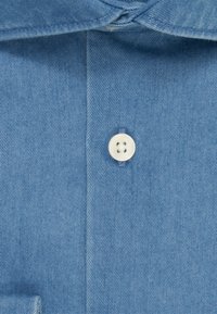 Camicia di denim azzurro leggero con colletto classico, un singolo bottone bianco e un tessuto testurizzato con sottili motivi diagonali.