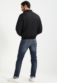 Zwarte gewatteerde bomberjack met geribde manchetten en kraag, gedragen met blauwe jeans en witte sneakers, gezien vanaf de achterkant.