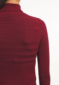 Maglione bordeaux con texture a coste e motivo a righe orizzontali; presenta un collo alto e una silhouette aderente. Materiale morbido.