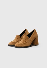 Mocassini da donna in suede marrone con tacco a blocco, suola nera e dettaglio a fascia sulla parte superiore, esposti su uno sfondo bianco semplice.