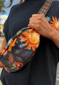 Värikäs kuviollinen olkalaukku, jossa auringon ja käden kuviot, oranssit yksityiskohdat ja metallinen koriste. Säädettävä hihna, kevyt kangas.