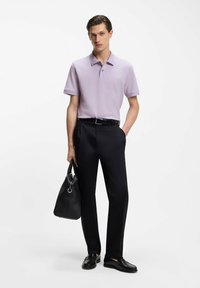 Fioletowa koszulka polo z krótkim rękawem, czarne spodnie, czarne skórzane buty, trzyma czarną skórzaną torbę z srebrnymi okuciami, stoi na tle białego tła.