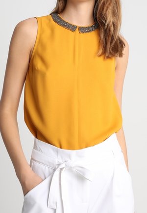 Mouwloze mosterdgele blouse met een kralen kraag, gecombineerd met witte broek met hoge taille, gestrikt met een stoffen ceintuur.