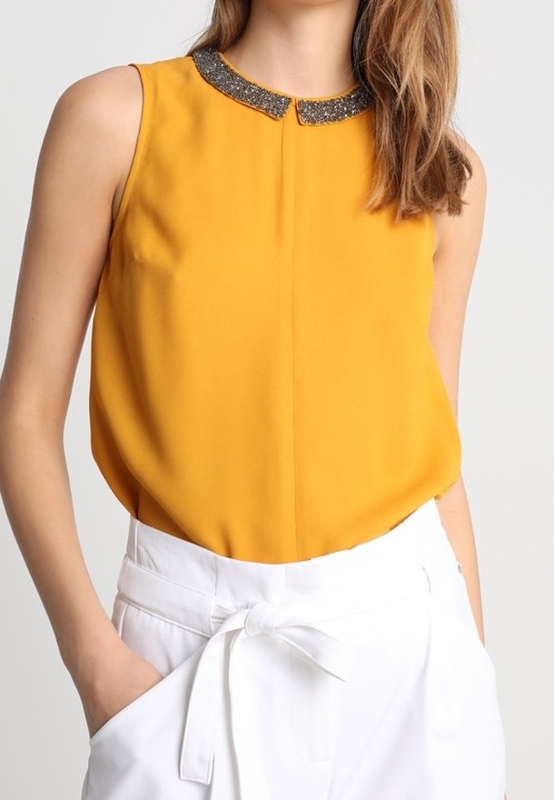 Blouse sans manches jaune moutarde avec un col orné de perles, associée à un pantalon blanc taille haute noué avec une ceinture en tissu.