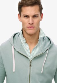 Hoodie in lichtgroene stof met een voorkantrits en lichtroze trekkoorden, gedragen over een blauw-wit gestreept shirt.