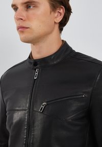 Zwarte leren jas met een hoge kraag, voorzien van een frontale rits en een klein ritsvak op de borst. Gladde textuur en slim-fit ontwerp.