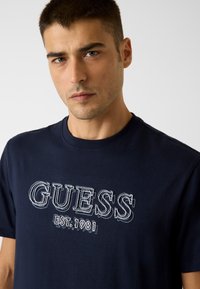 Marineblaues Baumwoll-T-Shirt mit einem großen, gestickten "GUESS"-Logo in kontrastierender Farbe, Rundhalsausschnitt und kurzen Ärmeln.