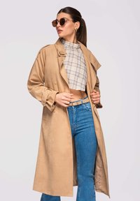 Femme portant un long trench-coat beige, un haut court à carreaux, un jean bleu, des lunettes de soleil et des boucles d'oreilles, posant devant un fond clair.