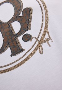 Witte stof met een groot, getextureerd, bruin logo met een luipaardprintpatroon, versierd met metalen studs en accenten.