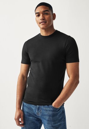 Young man wearing a fitted black short-sleeve shirt and blue jeans, standing with one hand in pocket against a plain background.