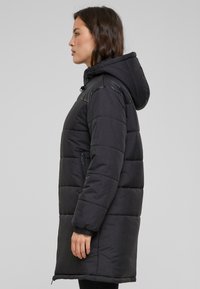 Abrigo de invierno negro acolchado con capucha, que presenta secciones acolchadas, puños elásticos y bolsillos laterales con cremallera. Textura suave con un acabado mate.
