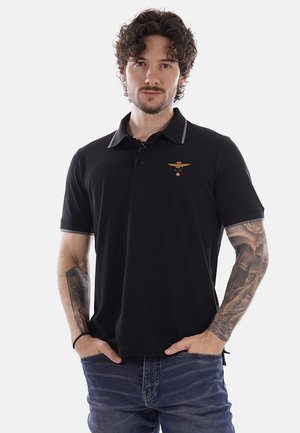 Uomo con capelli ricci che indossa una polo nera con un piccolo emblema dorato e jeans blu aderenti, mani nelle tasche, tatuaggi visibili sulle braccia.