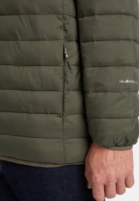 Primer plano de una persona que lleva una chaqueta acolchada verde oliva con cremallera visible y el logo de "Quiksilver" en la manga, combinada con jeans de color azul oscuro.