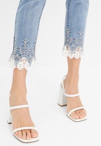Světle modré zkrácené džíny s bílou krajkovou květinovou výšivkou na lemu, doplněné bílými sandály na blokovém podpatku s tenkými pásky.