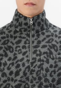 Hallhalli hallide musta leopardimustriga fleece-pullover, kõrge kaelusega ja pool tõmblukuga. Omab esi-taskut ja tekstuurset kangast.