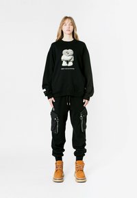 Sudadera negra de gran tamaño con estampado gráfico, combinada con pantalones cargo negros que tienen bolsillos laterales y cremalleras, complementados con botas marrón.
