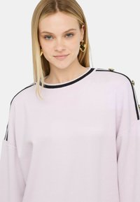 Pullover rosa chiaro con scollatura rotonda, strisce orizzontali nere sulle spalle e dettagli in bottone dorato ai lati. Tessuto morbido e testurizzato.
