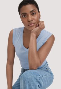 Helles blaues ärmelloses Oberteil mit V-Ausschnitt, aus weichem Stoff. Kombiniert mit hochgeschnittenen blauen Jeans und einem neutralen Hintergrund.