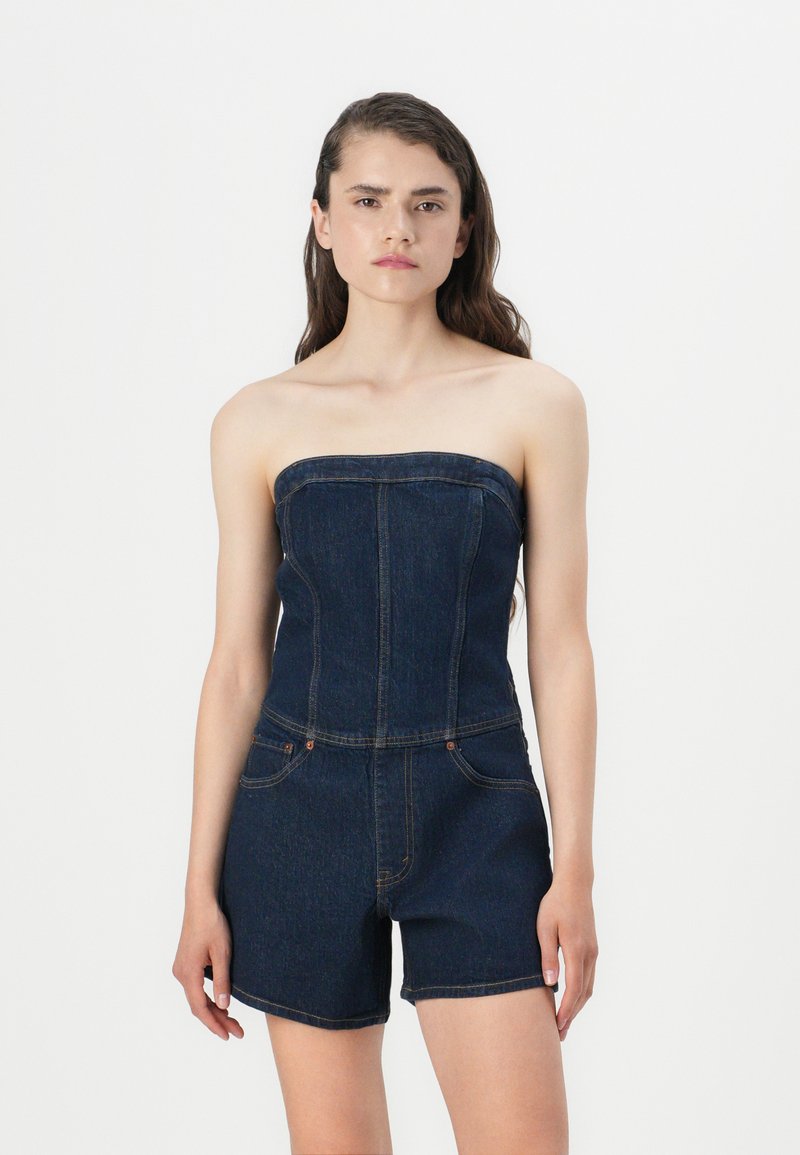 Dongeribukse uten stropper i mørkeblått, med en strukturert overdel, tettsittende design og korte shorts, med oransje sting og lommer.