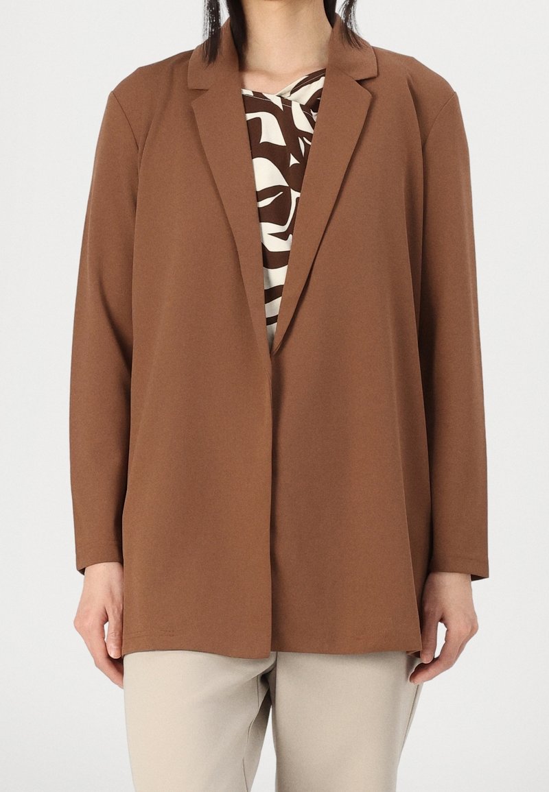 Brown blazer with a smooth texture, featuring a notched collar and single button closure, worn over a patterned top and beige pants.