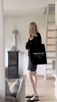 Zwarte tote bag met witte tekst "ALLSAINTS" opvallend weergegeven. Grijze jurk, zwarte schoenen en een moderne inrichting met een metalen fornuis.