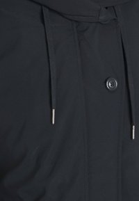 Mustan takin, jossa on nappikiinnitys, säädettävä naruhuppu ja sileä, matta tekstuuri hienovaraisilla ompeluyksityiskohdilla.