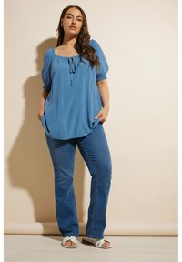 Blå, löst sittande topp med korta, puffiga ärmar och knytning fram. Kombinerad med åtsittande blå jeans och vita, remmade sandaler.