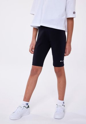 Pantaloni shorts neri in tessuto elasticizzato, caratterizzati da una vestibilità aderente, abbinati a una t-shirt bianca ampia e a sneakers bianche con dettagli verdi.