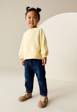 Jaunas vaikas dėvi šviesiai geltoną megztinį, tamsiai mėlynas džinsus ir leopardžio rašto sportbačius, stovi neutraliame fone.