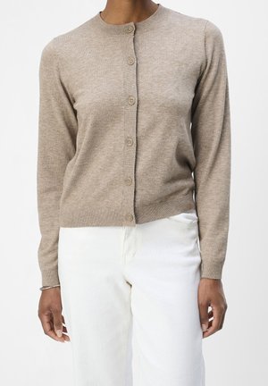 Cardigan beige ajusté à boutons porté par une personne en pantalon blanc, debout sur un fond blanc uni.