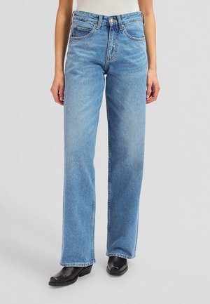 Person iført lyseblå høy på livet, rette jeans og svarte støvler med firkantet tå mot en ensfarget grå bakgrunn.