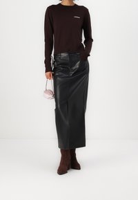 Frau in einem dunkelbraunen Pullover, schwarzer Leder-Midikleid, braunen Wildlederstiefeln, die eine kleine hellrosa Handtasche hält, vor einem weißen Hintergrund stehend.