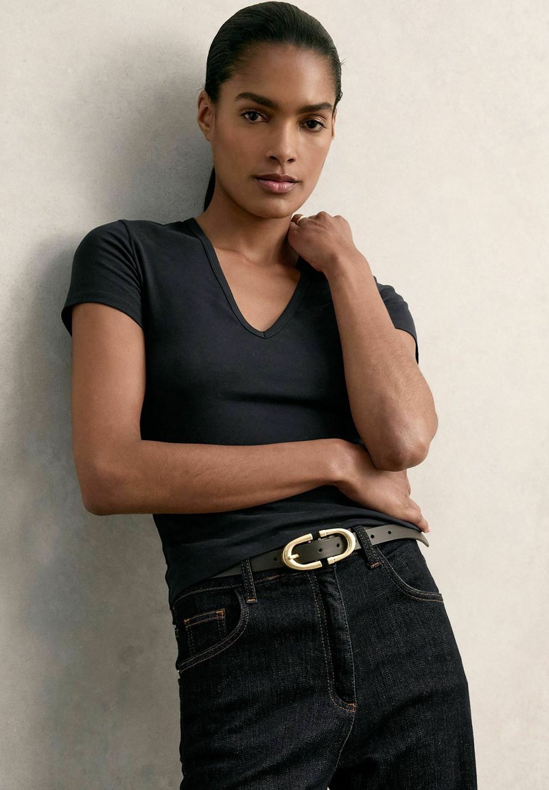 Tricou negru cu guler în V, fabricat din bumbac, purtat cu blugi denim închisi și o centură neagră cu o cataramă ovală aurie. Textură netedă și design ajustat.