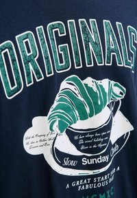 Mørkeblå t-shirt med fed, slidt hvid og grøn tekst. Har et grafik af en croissant på en kop, samt yderligere tekst nedenfor.