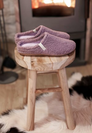 Een paar paarse pluizige pantoffels op een houten krukje naast een brandende open haard en een zwart-witte bontkleed op de houten vloer.