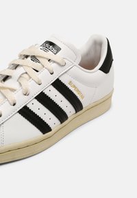 Vit läder sneakers med svarta ränder, texturerade detaljer och beige sula. Har krämfärgade snören och "SUPERSTAR"-logotyp i guld.
